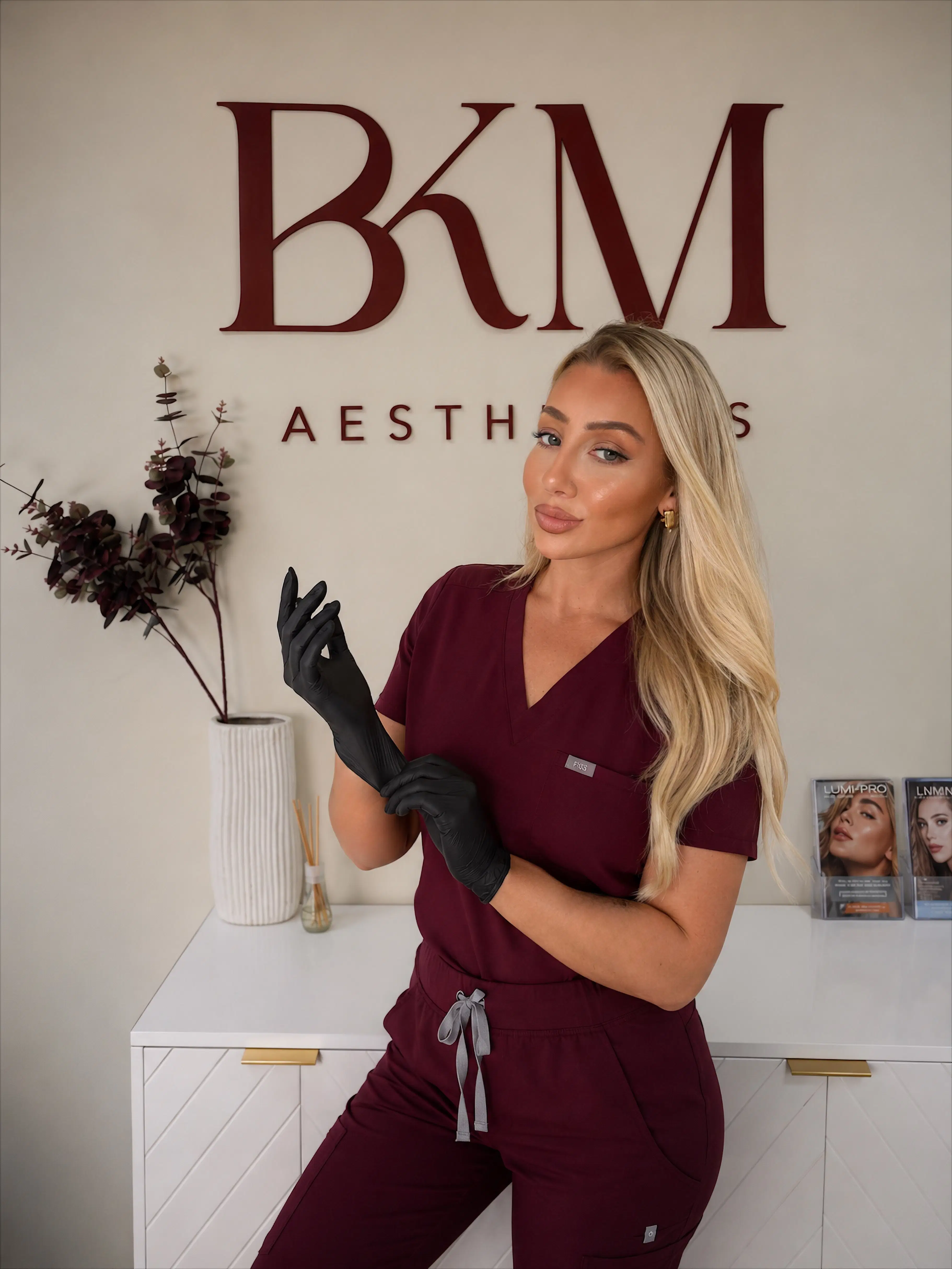 Buckingham Aesthetics practitioner in burgundy scrubs and black gloves, standing in front of the clinic signage